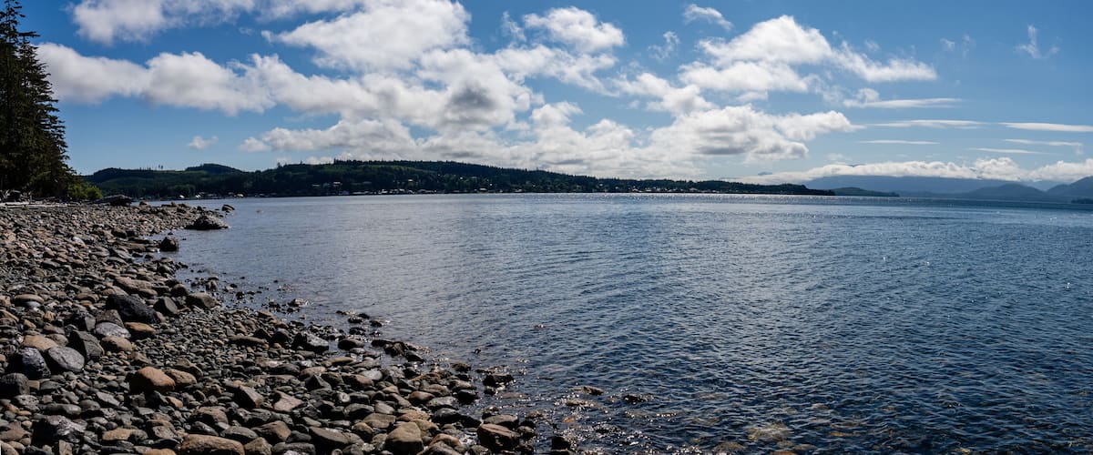 A rocky shoreline with calm water, a clear sky, and distant hills.