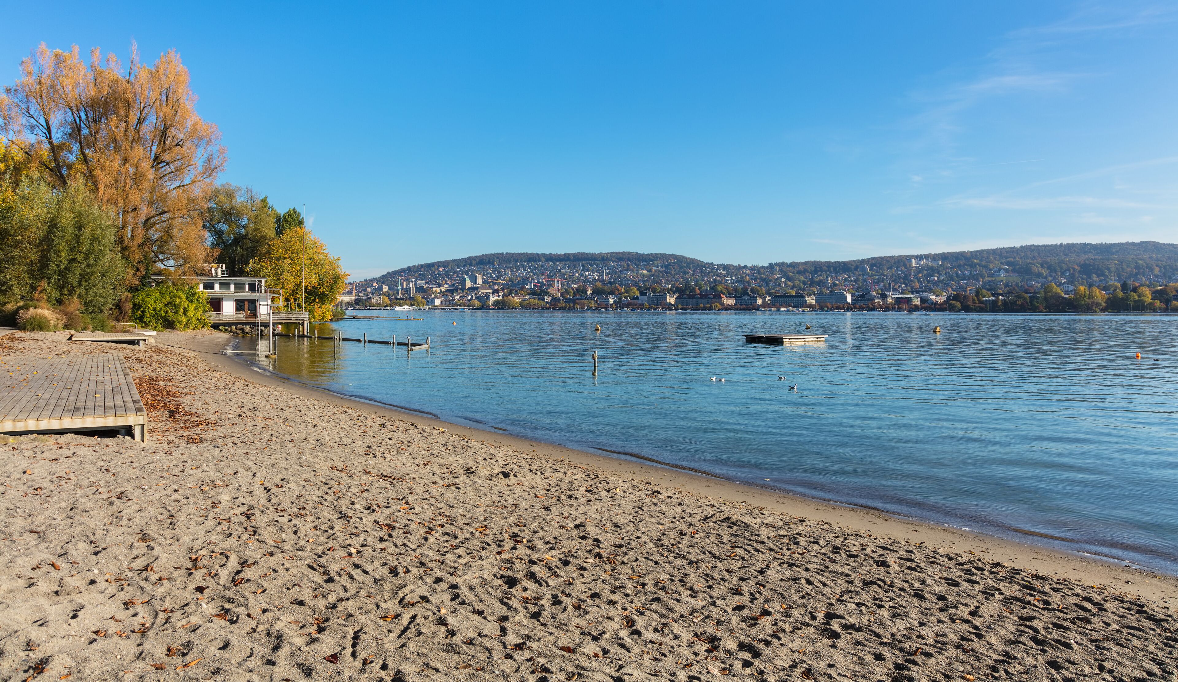 Una playa arenosa con un muelle de madera, agua tranquila y un paisaje urbano al fondo.