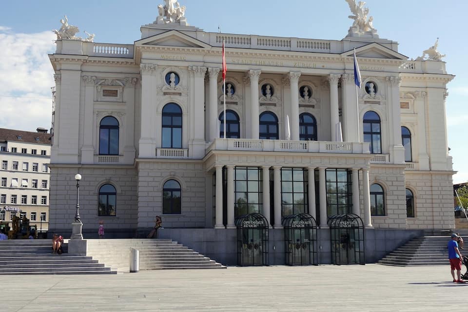 A grand classical building with columns and statues, featuring the text "Stadt Theater Zurich" and "Opernhaus Zurich" on its facade.