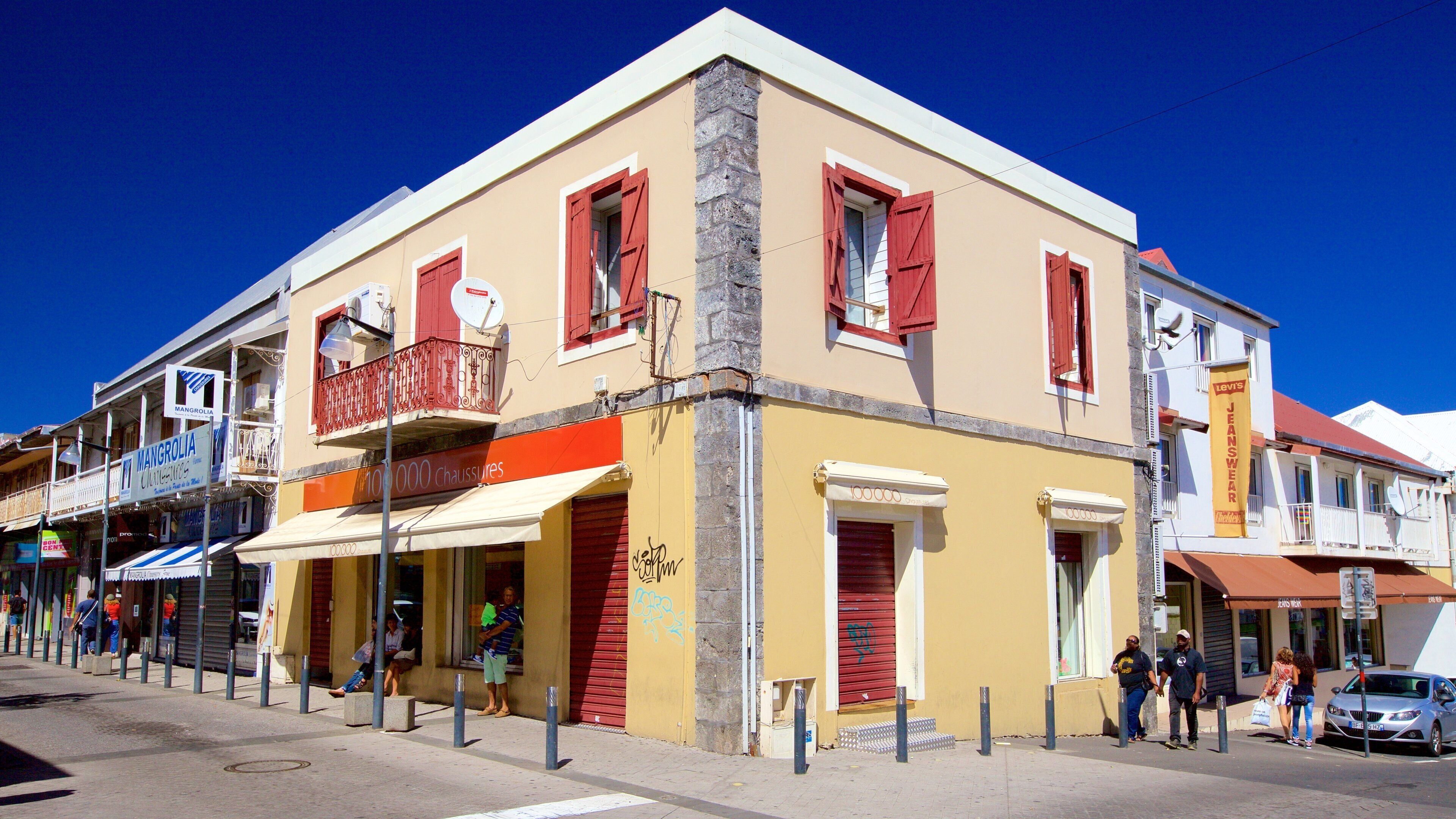 A yellow building with red shutters and a balcony, storefronts, and pedestrians.