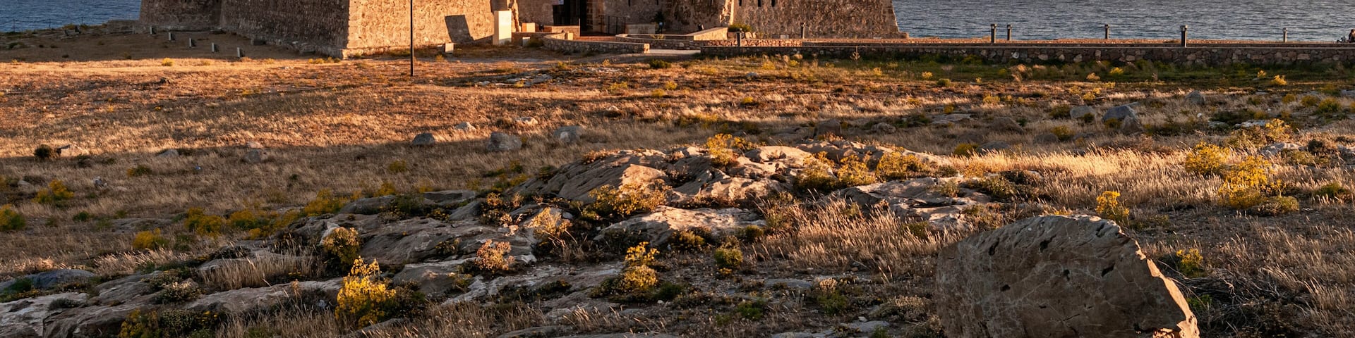 A coastal fortification with a large stone building, a rocky foreground, and the sea in the background.