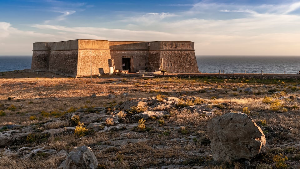 A coastal fortification with a large stone building, a rocky foreground, and the sea in the background.