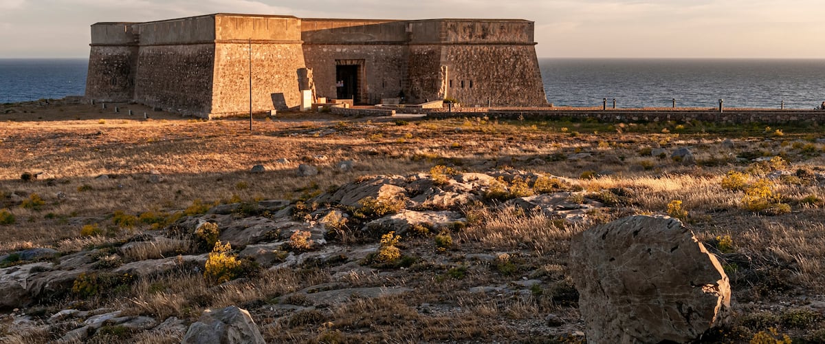 A coastal fortification with a large stone building, a rocky foreground, and the sea in the background.
