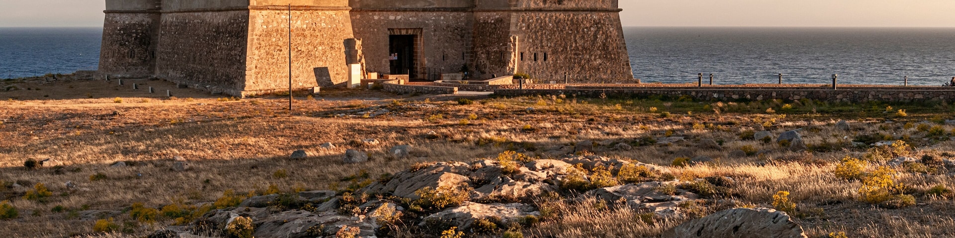 A coastal fortification with a large stone building, a rocky foreground, and the sea in the background.