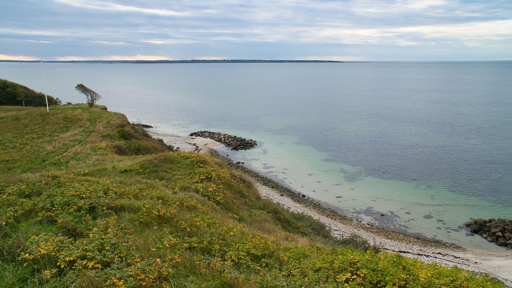 Eine Küstenlandschaft mit einem grasbewachsenen Hügel, einer steinigen Küstenlinie und einem ruhigen Meer unter einem bewölkten Himmel.