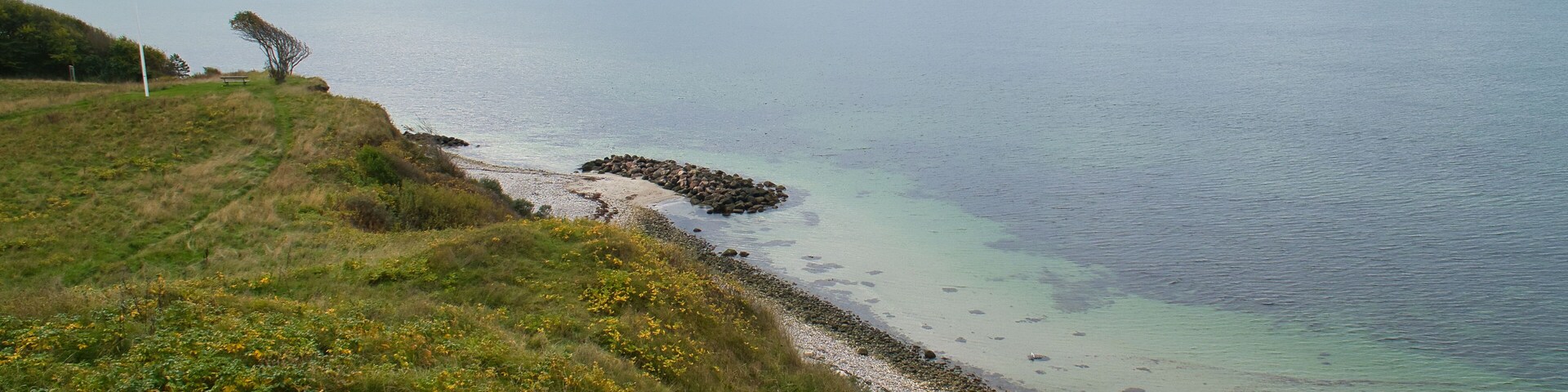 A coastal landscape with a grassy hill, a rocky shoreline, and a calm sea under a cloudy sky.