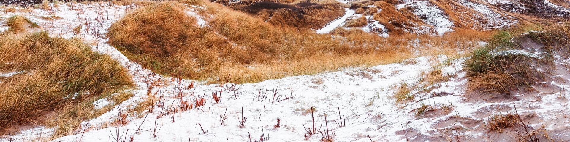A white lighthouse on a sandy hill with dry grass and snow patches, under a cloudy sky.