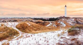 A white lighthouse on a sandy hill with dry grass and snow patches, under a cloudy sky.