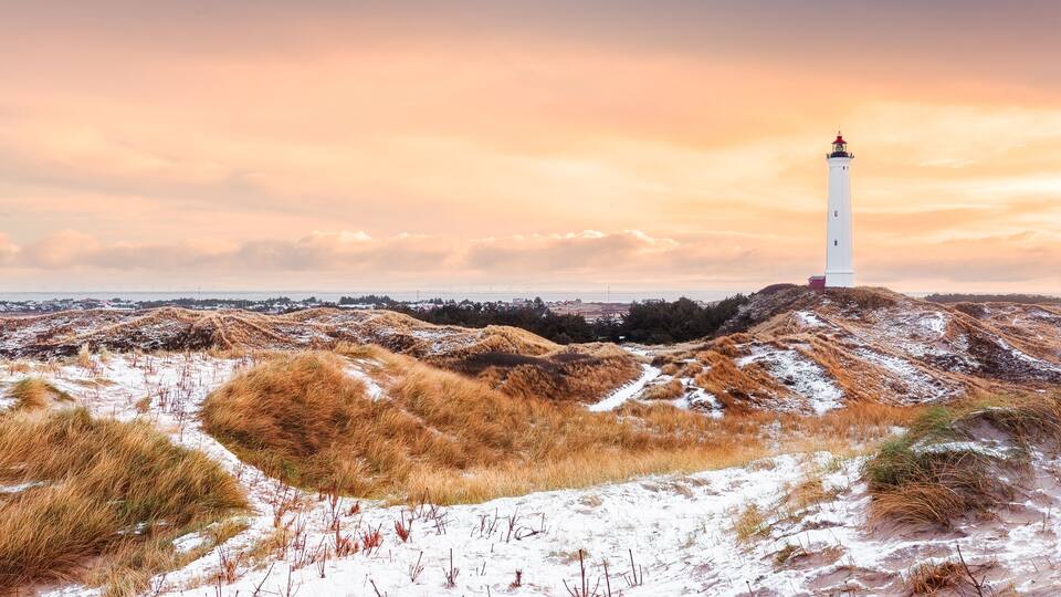 Un faro blanco sobre una colina arenosa con pasto seco y parches de nieve, bajo un cielo nublado.
