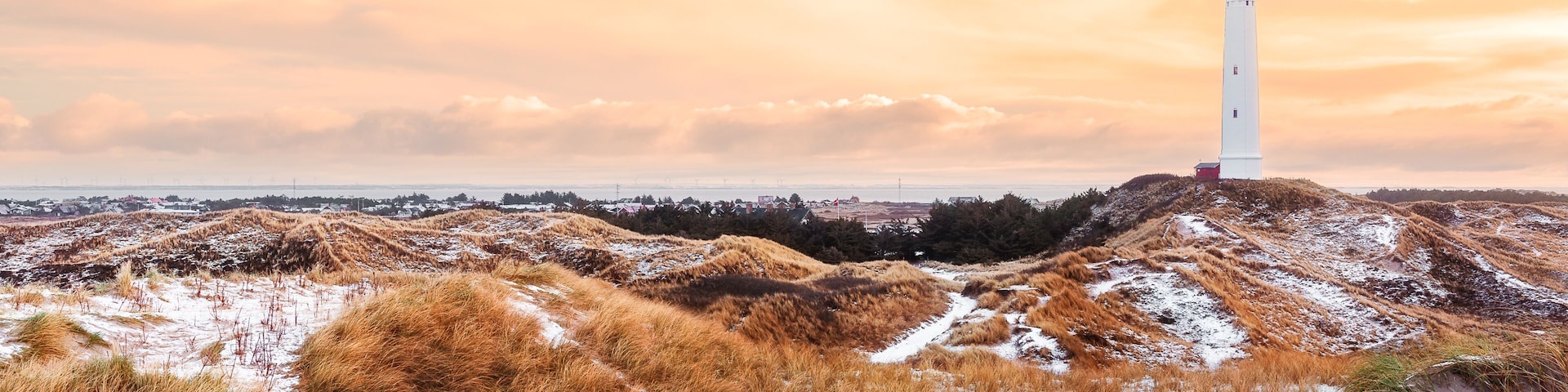 A white lighthouse on a sandy hill with dry grass and snow patches, under a cloudy sky.