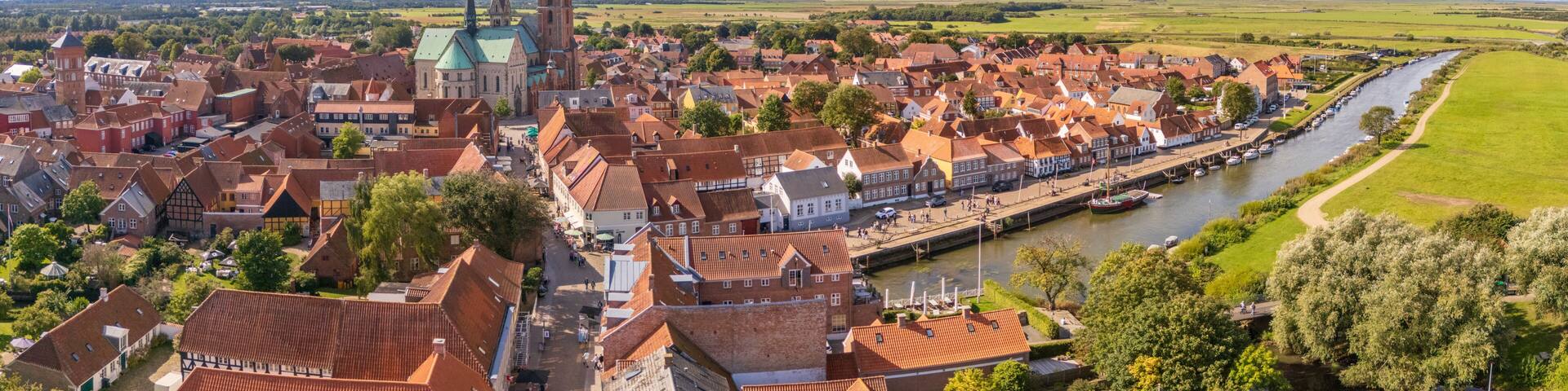 Aerial view of the serene Ribe Cathedral towering over the town's red rooftops, nestled beside a tranquil river reflecting the sky, Ribe, Denmark.