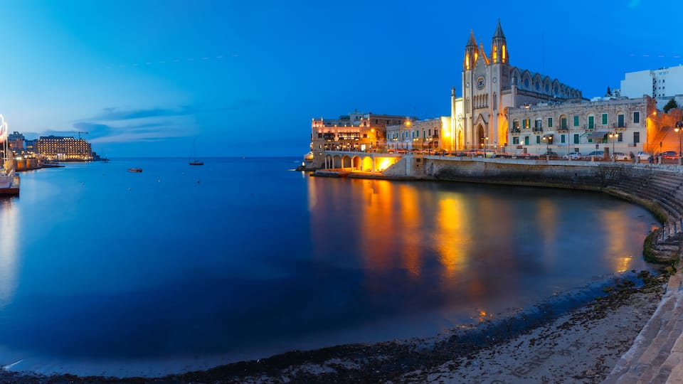 A waterfront cityscape with a prominent church, calm sea, and illuminated buildings.