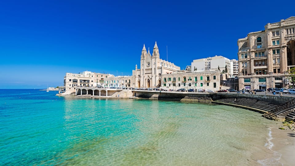 A coastal cityscape with a prominent church, clear turquoise waters, and a sandy beach.