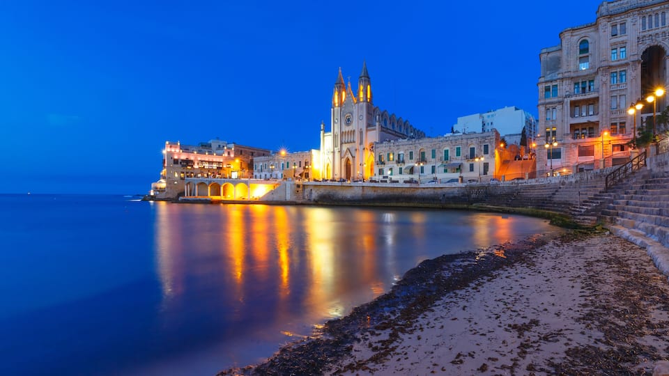 A waterfront cityscape with illuminated buildings, a prominent church with twin towers, and a calm sea reflecting the lights.