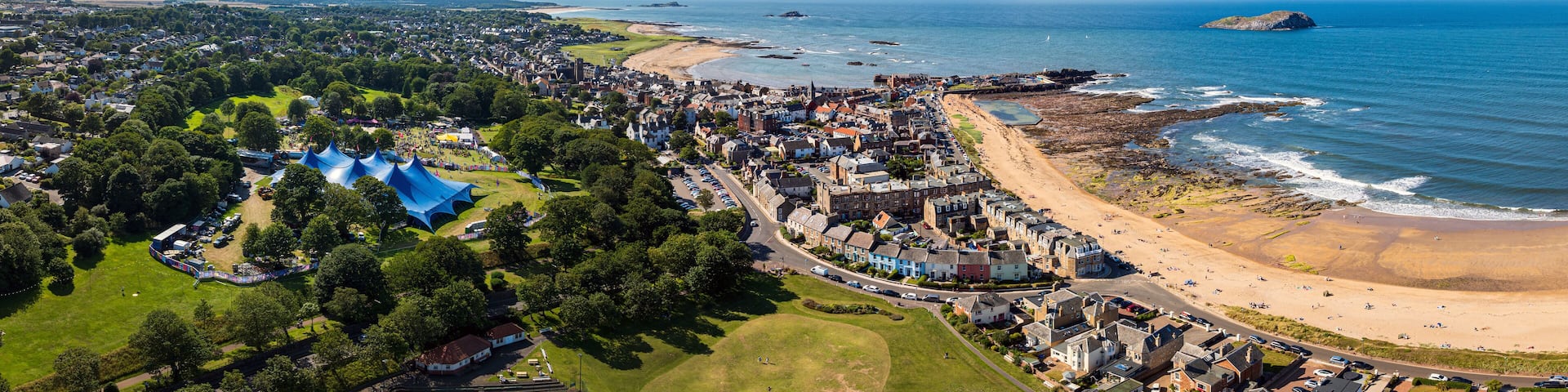 Aerial view of a coastal town where the vibrant blue of a large tent contrasts with the golden sands and azure sea, North Berwick, Scotland, United Kingdom.