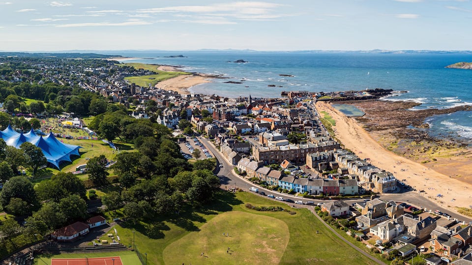 Aerial view of a coastal town where the vibrant blue of a large tent contrasts with the golden sands and azure sea, North Berwick, Scotland, United Kingdom.