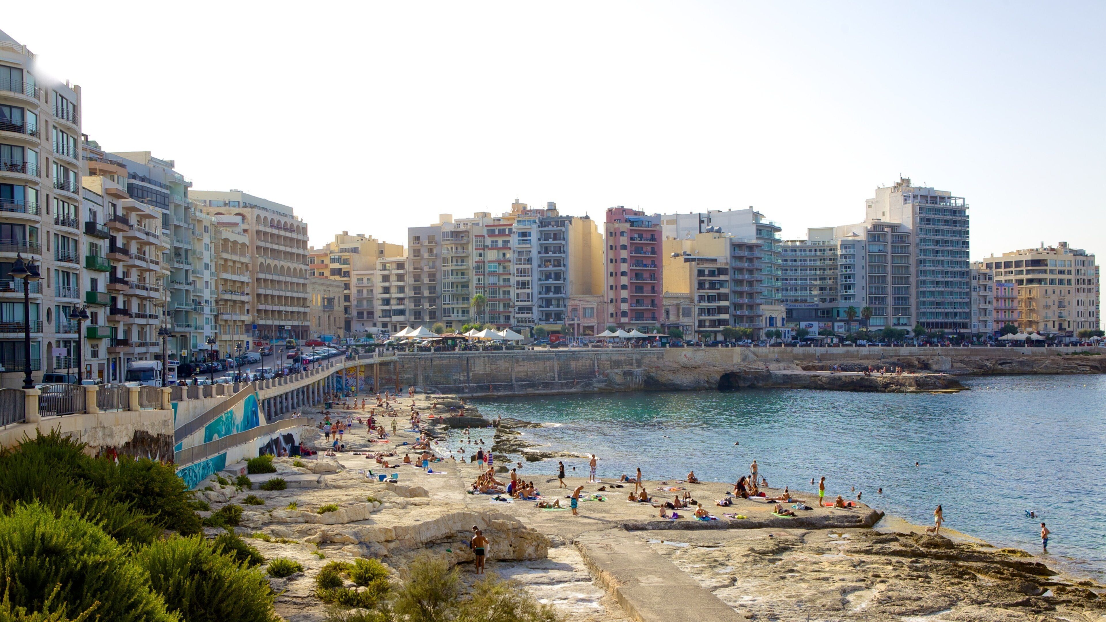 A coastal cityscape with a rocky beach, people sunbathing, and multi-story buildings in the background.