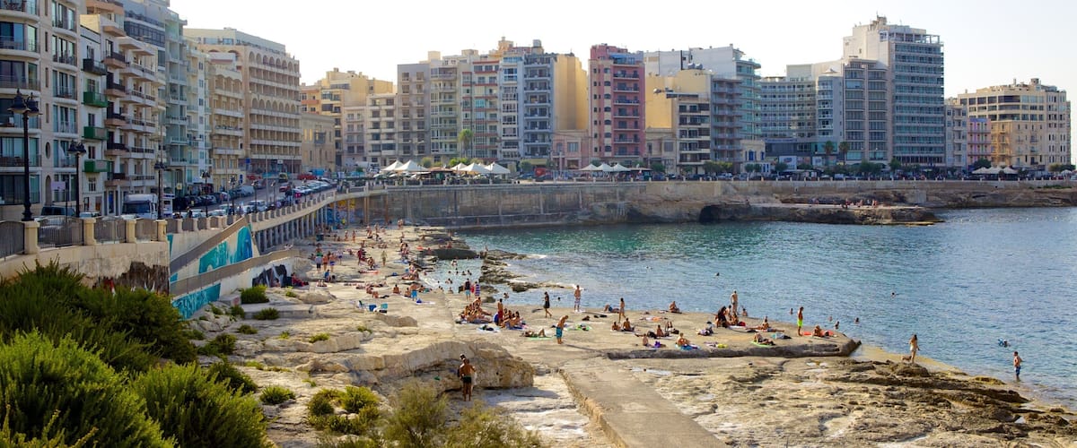 A coastal cityscape with a rocky beach, people sunbathing, and multi-story buildings in the background.