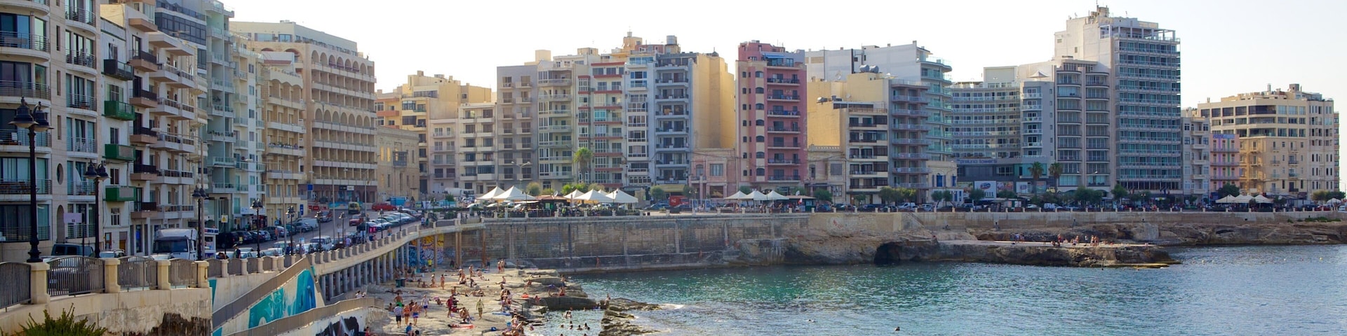 A coastal cityscape with a rocky beach, people sunbathing, and multi-story buildings in the background.