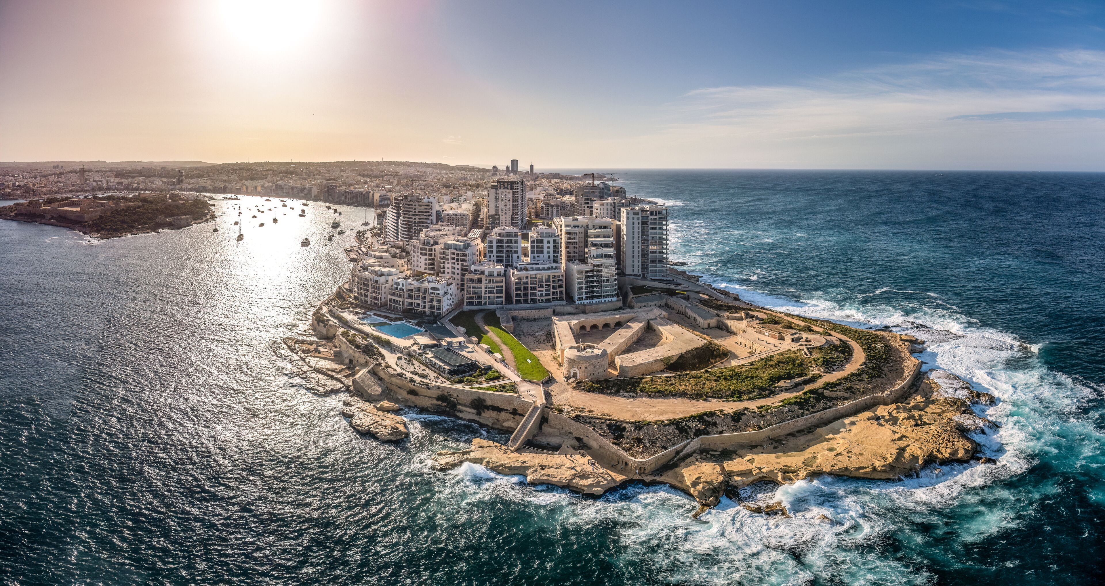 A coastal cityscape with a prominent fortress-like structure surrounded by modern buildings, a marina with boats, and a clear view of the ocean.