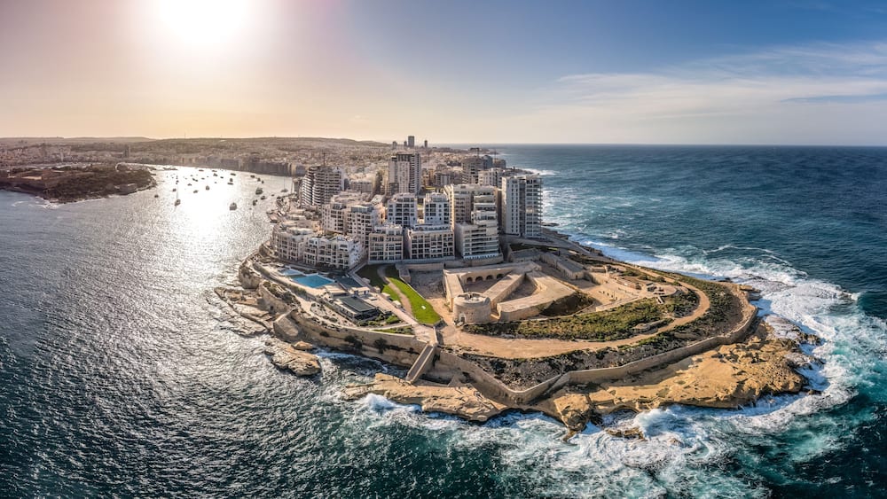 A coastal cityscape with a prominent fortress-like structure surrounded by modern buildings, a marina with boats, and a clear view of the ocean.