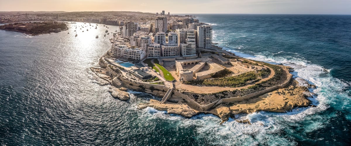 A coastal cityscape with a prominent fortress-like structure surrounded by modern buildings, a marina with boats, and a clear view of the ocean.