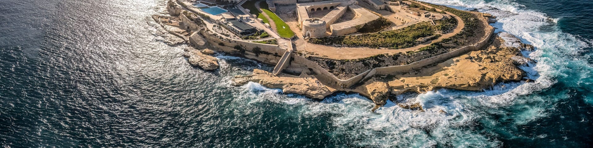 A coastal cityscape with a prominent fortress-like structure surrounded by modern buildings, a marina with boats, and a clear view of the ocean.