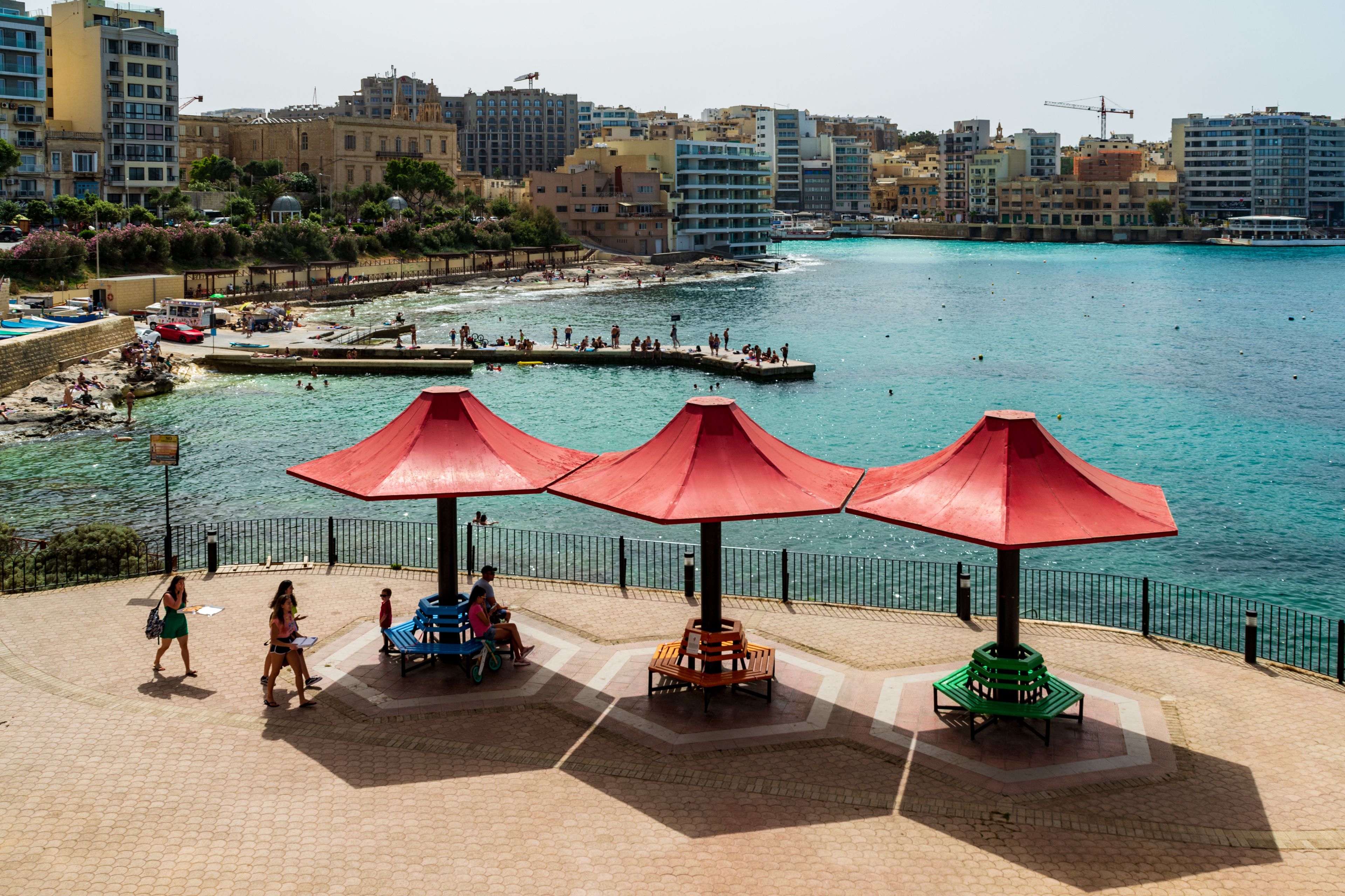 Un paseo marítimo costero con sombrillas rojas, una playa de arena y un paisaje urbano al fondo.
