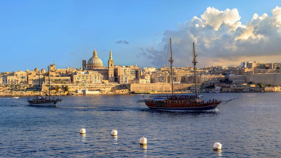 A coastal cityscape with historic buildings, including a prominent dome and a fortified wall, and two boats on the water.