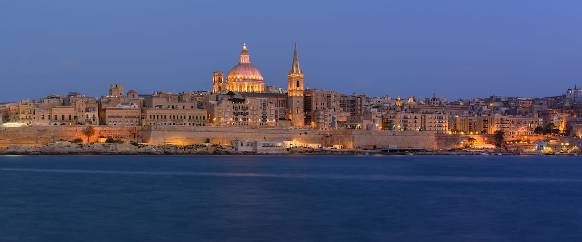 A coastal cityscape at dusk with a prominent domed cathedral and a tall minaret.