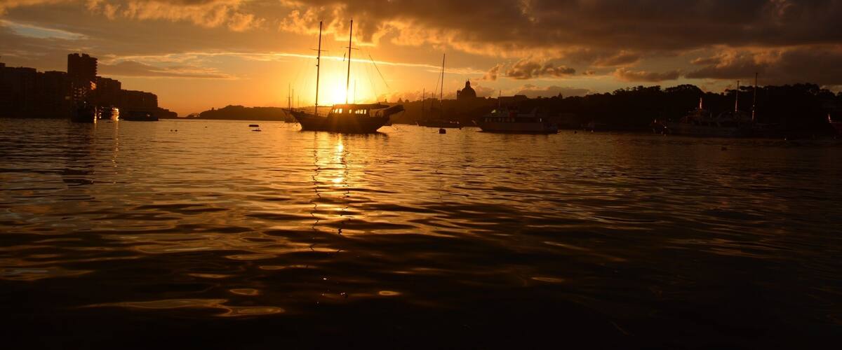 Un coucher de soleil sur un port, avec des bateaux et des bâtiments se détachant en silhouette sur le ciel.