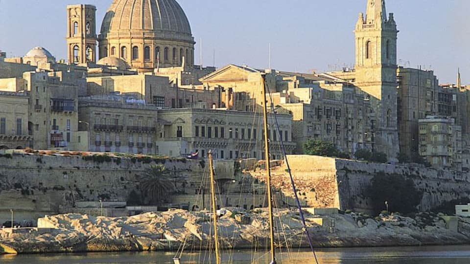A coastal cityscape with a prominent dome and a tall tower. A sailboat is anchored in the foreground.