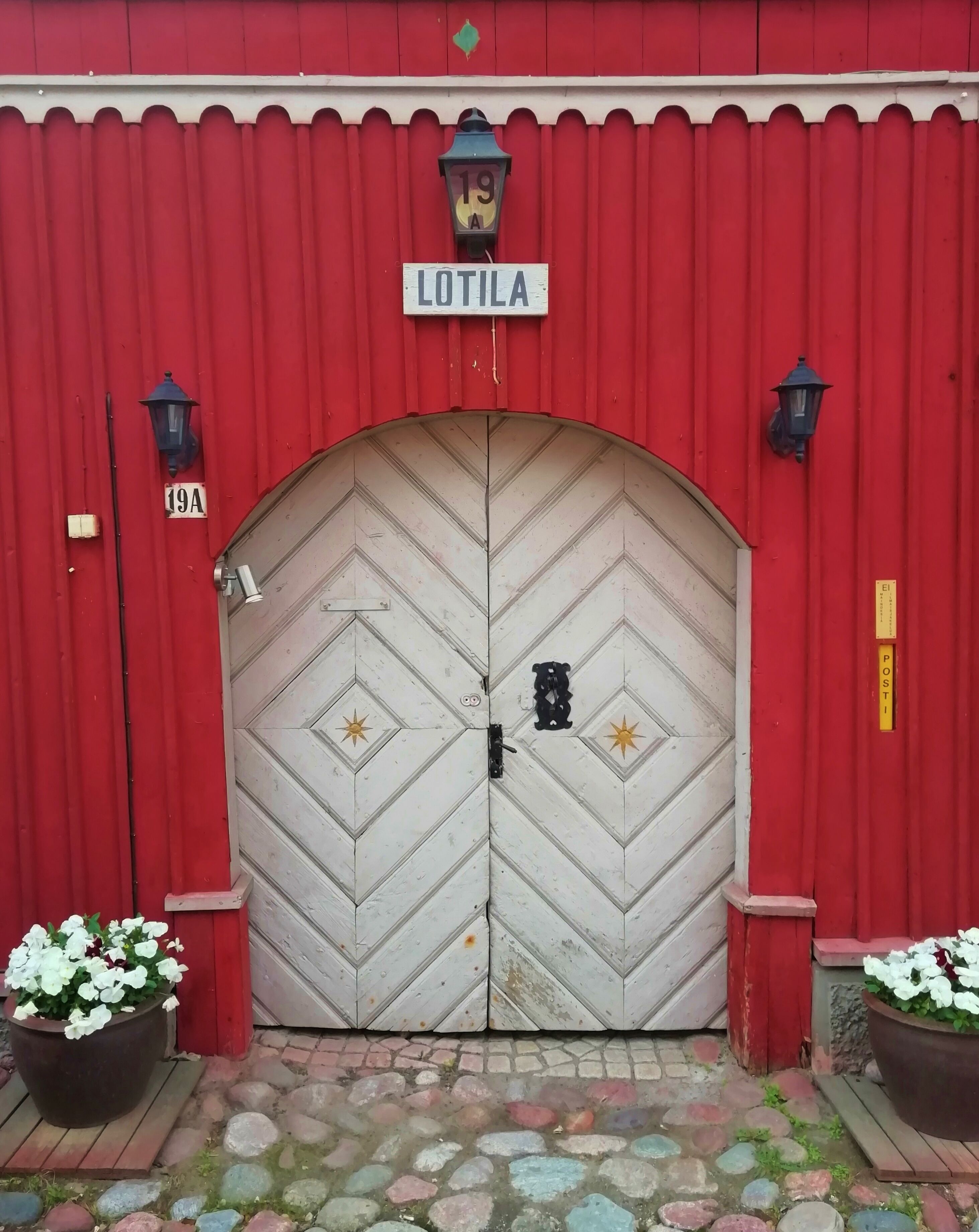 Punainen rakennus, jossa on valkoinen ovi, ja sen yläpuolella on kyltti, jossa lukee ”LOTILA”. Lisäksi siinä on kaksi ruukussa olevaa kasvia, joissa on valkoiset kukat.