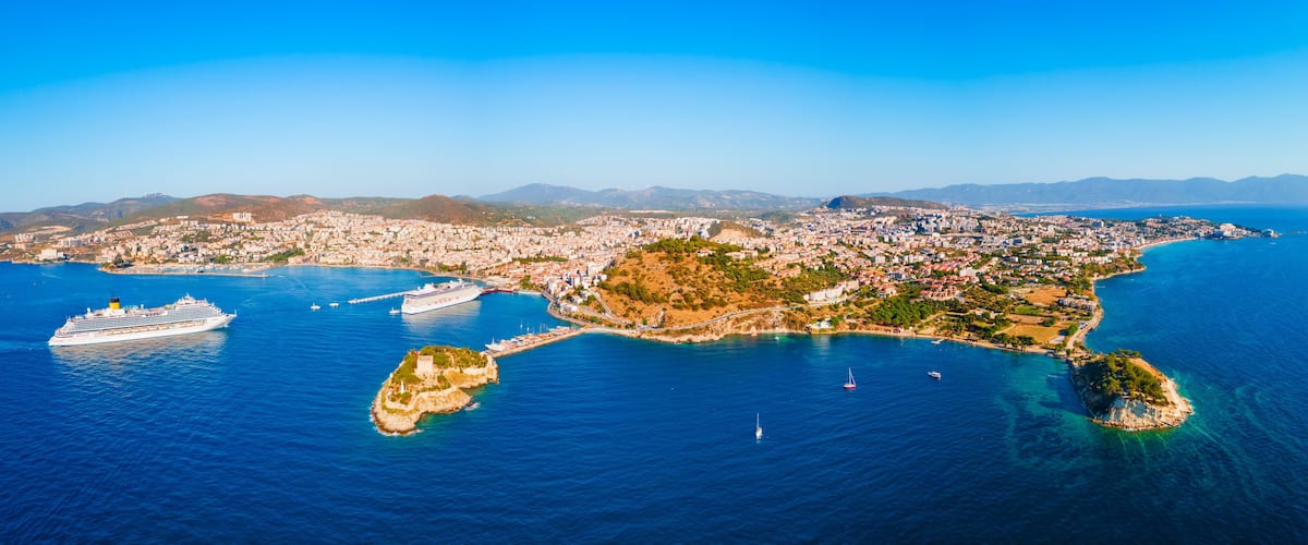 A coastal cityscape with a cruise ship docked near the shore, surrounded by clear blue waters and a mountainous backdrop.