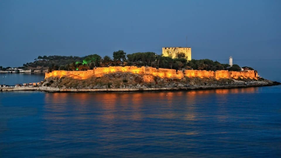 A fortified island with a prominent tower and lighthouse, surrounded by calm water and illuminated by artificial lighting.