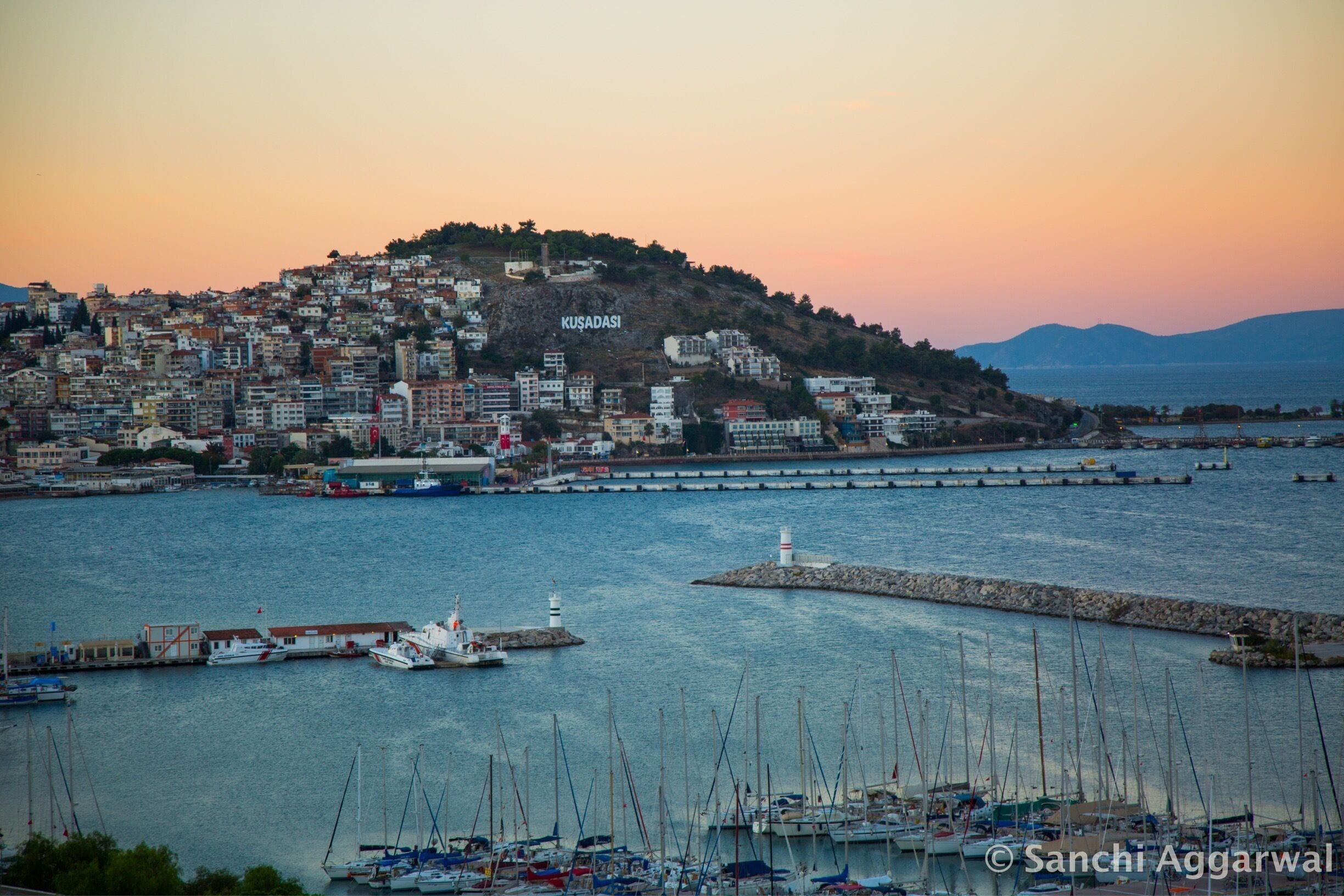 A coastal cityscape with a prominent hill labeled "KUSADASI" and a marina with numerous boats.