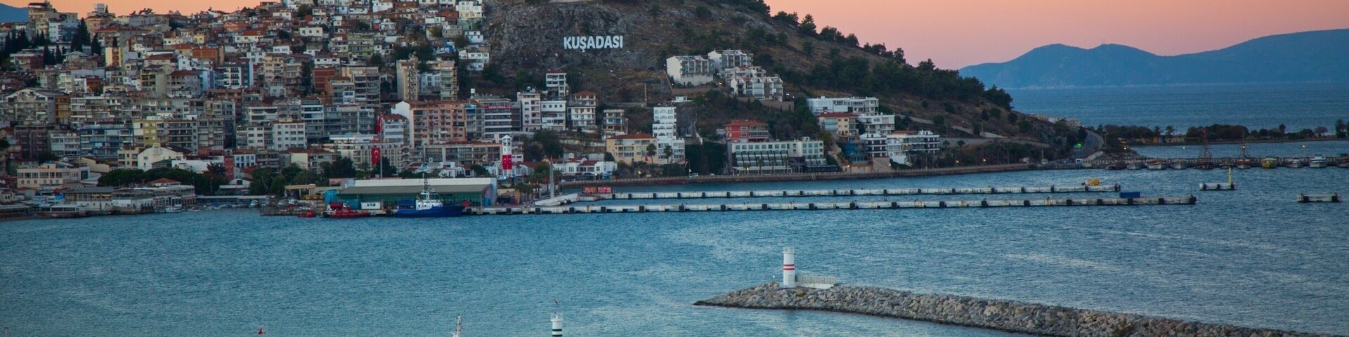 A coastal cityscape with a prominent hill labeled "KUSADASI" and a marina with numerous boats.