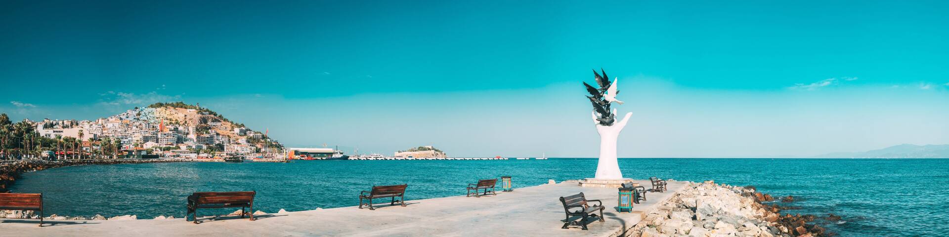 A coastal cityscape with a prominent white statue, benches, and a rocky shoreline.