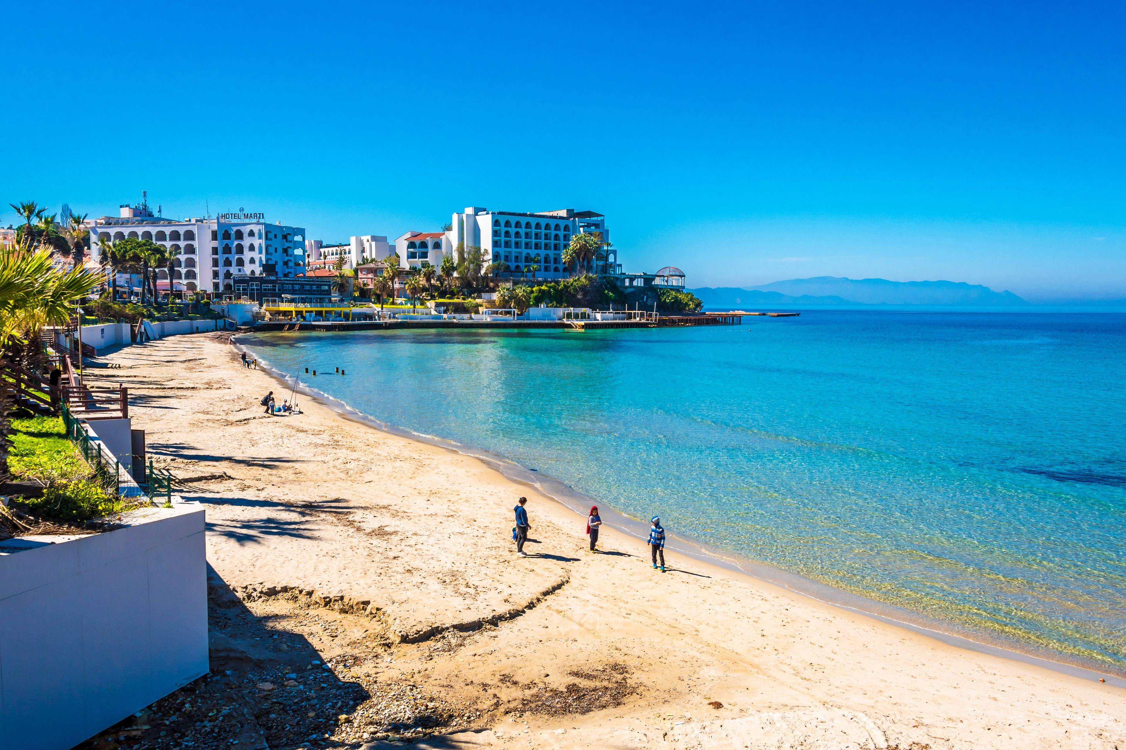 Una playa soleada con aguas turquesas cristalinas, una orilla arenosa y edificios al fondo.