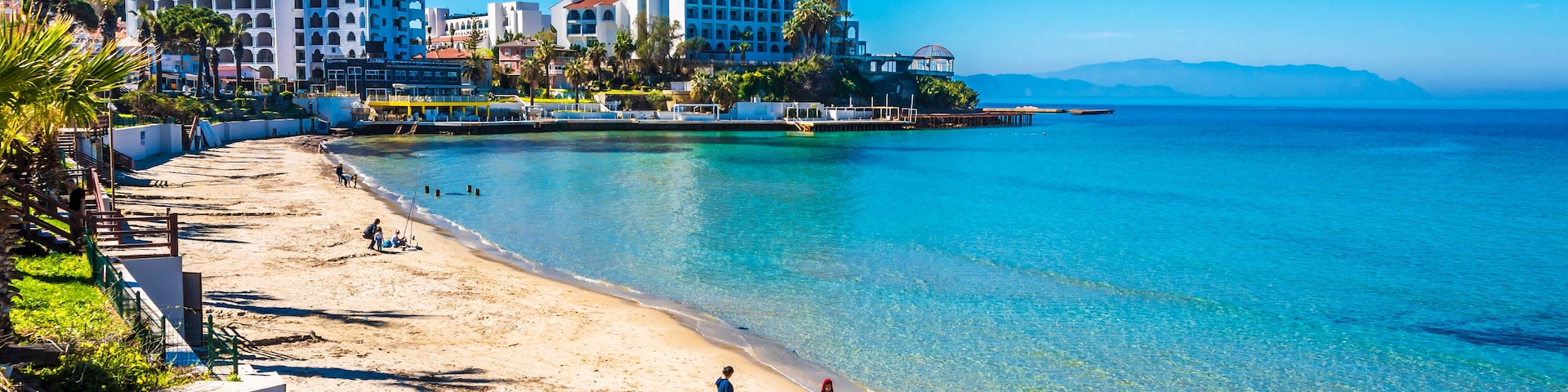 Una playa soleada con aguas turquesas cristalinas, una orilla arenosa y edificios al fondo.
