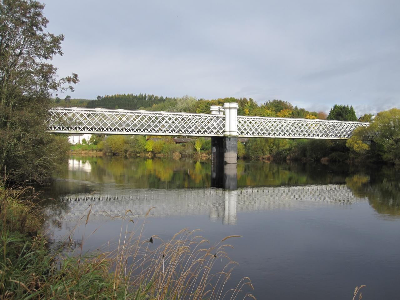 A white lattice bridge over a calm river with surrounding greenery and trees.
