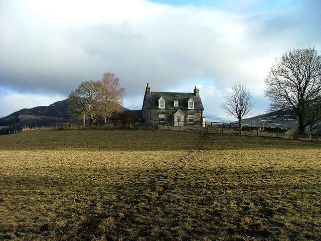 A large stone house with a green roof, surrounded by a grassy field and a few trees.