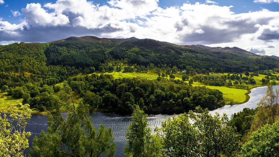 A lush valley with a river, dense forests, and rolling hills under a partly cloudy sky.
