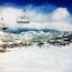 A snow-covered ski slope with ski lifts, buildings, and mountains in the background.