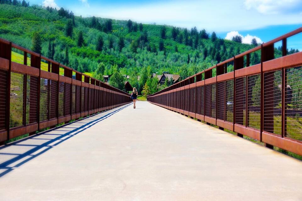 A pedestrian bridge with metal railings, leading to a forested hillside.