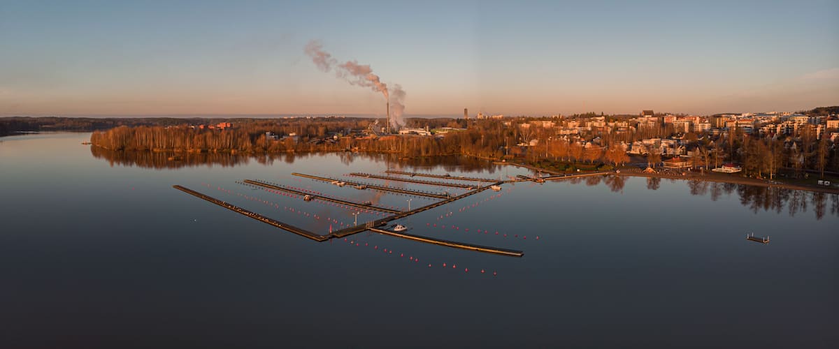 A calm lake with a dock, a smokestack, and a cityscape in the background.