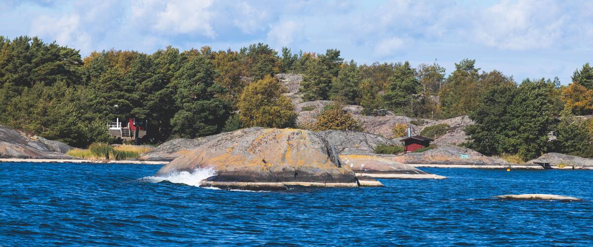 A rocky shoreline with a small red cabin, dense forest, and a clear blue sky.