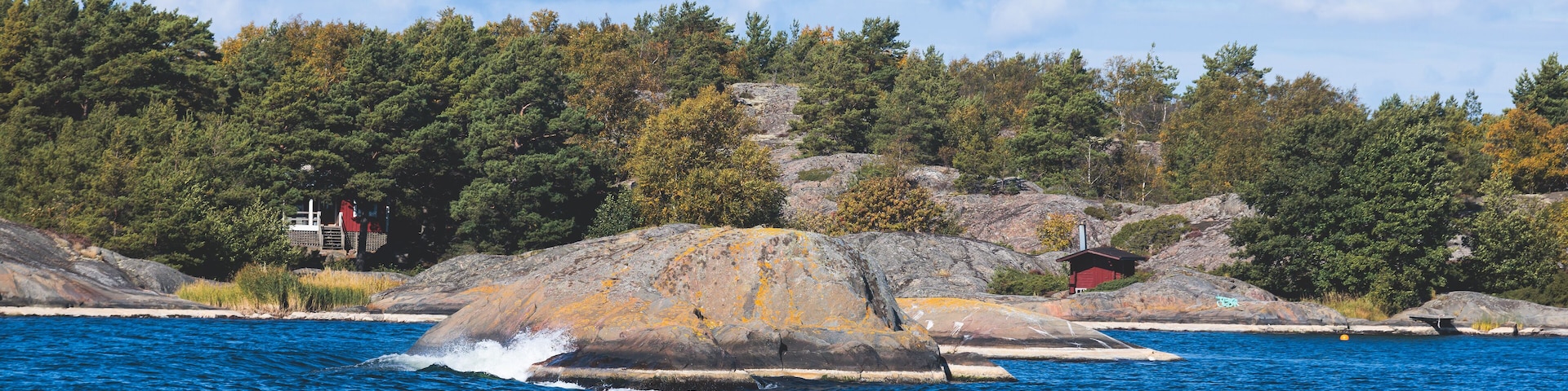 A rocky shoreline with a small red cabin, dense forest, and a clear blue sky.
