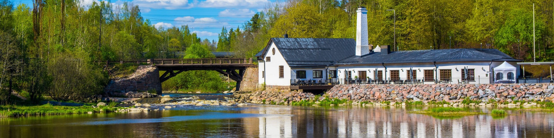 A riverside building with a bridge, surrounded by trees and a clear sky.