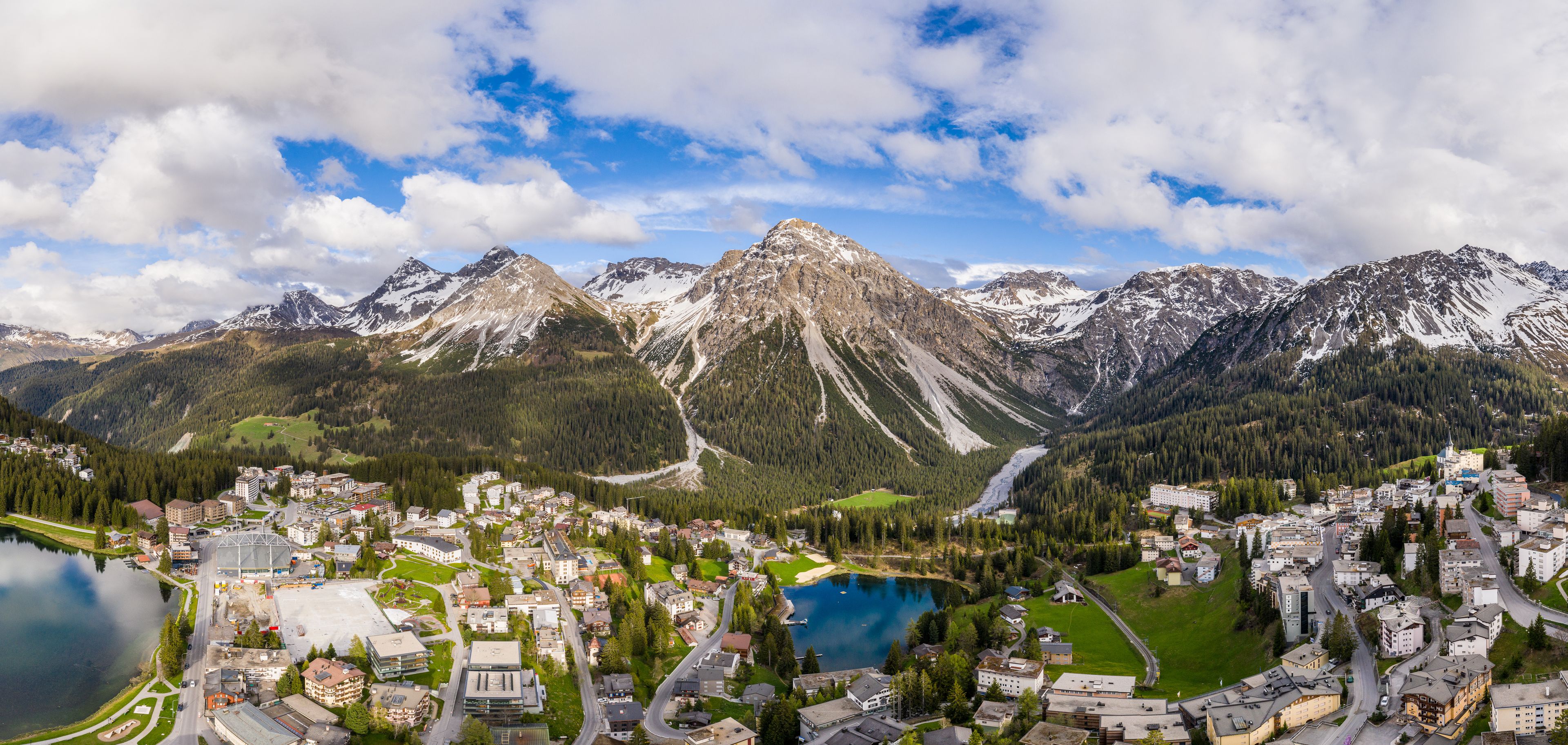 A mountainous landscape with a town nestled at the base, a lake, and snow-capped peaks in the background.
